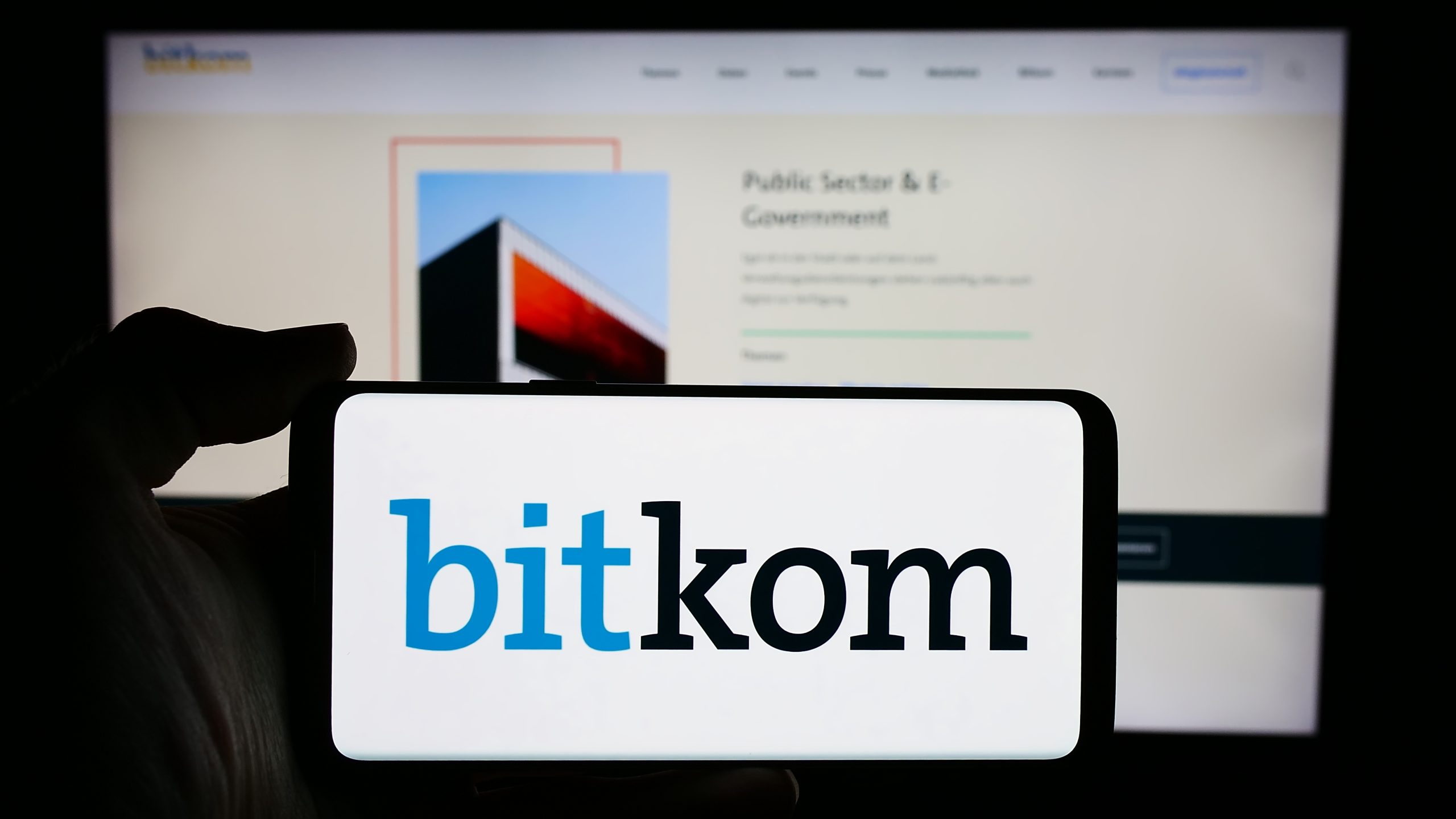 Eine Hand hält ein Telefon mit dem Bitkom-Logo vor einem verschwommenen Computerbildschirm, der eine Bitkom-Webseite zeigt.