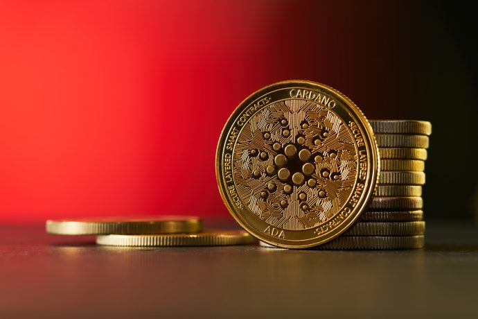 Ein Stapel goldfarbener Cardano (ADA) Kryptowährungsmünzen ist auf einer flachen Oberfläche angeordnet und hebt die Eleganz und Innovation von Cardano vor einem verschwommenen roten Hintergrund hervor.