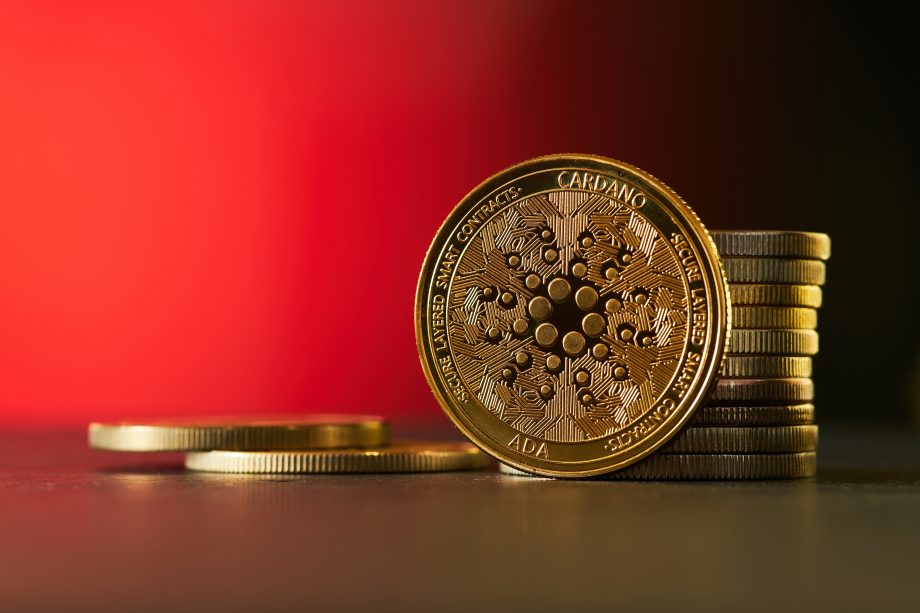 Ein Stapel goldfarbener Cardano (ADA) Kryptowährungsmünzen ist auf einer flachen Oberfläche angeordnet und hebt die Eleganz und Innovation von Cardano vor einem verschwommenen roten Hintergrund hervor.