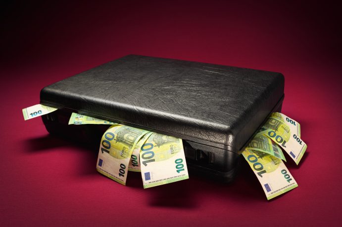 Eine geschlossene schwarze Aktentasche mit mehreren 100-Euro-Banknoten und Andeutungen von Krypto-Vermögenswerten, die vor einem dunkelroten Hintergrund herausragen.