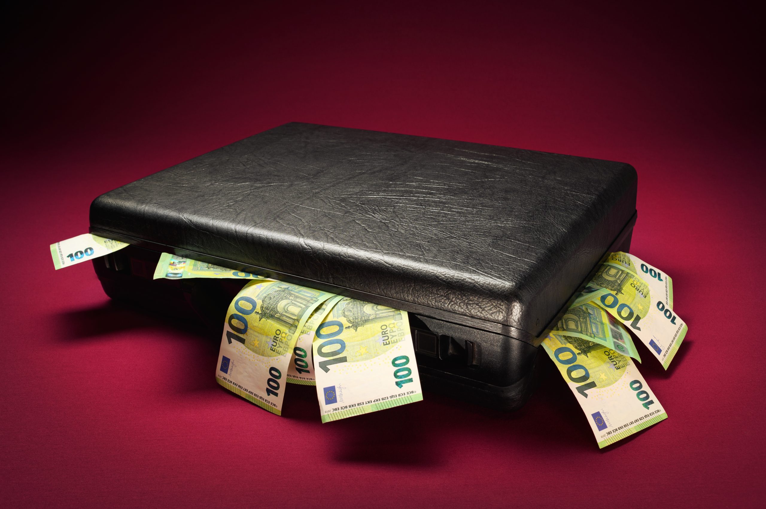 Eine geschlossene schwarze Aktentasche mit mehreren 100-Euro-Banknoten und Andeutungen von Krypto-Vermögenswerten, die vor einem dunkelroten Hintergrund herausragen.