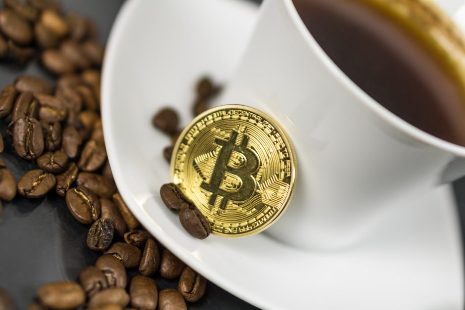 Eine goldene Bitcoin-Münze ruht auf einer weißen Kaffeetasse, die mit Kaffee gefüllt und von verstreuten Kaffeebohnen umgeben ist - eine stilvolle Anspielung auf die Welt der Kryptowährung.