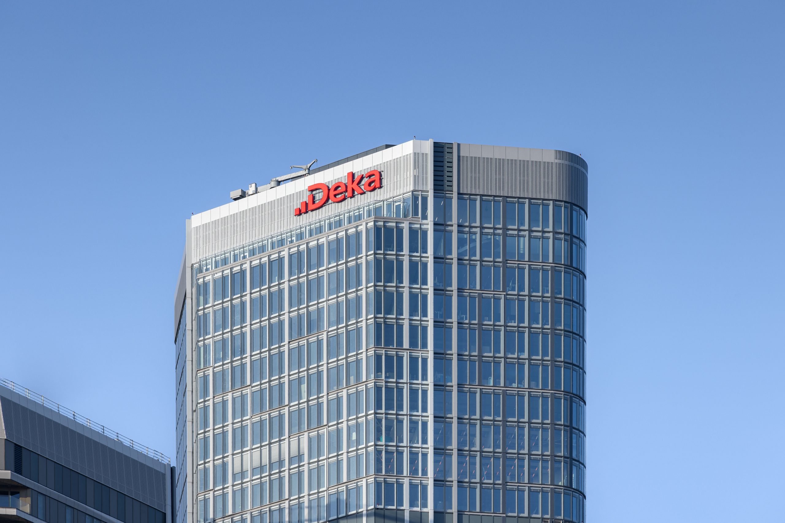 Ein modernes gläsernes Bürogebäude mit dem roten Deka-Logo an der Spitze steht vor einem strahlend blauen Himmel und symbolisiert Planungssicherheit, denn die Deka bereitet sich auf die Einführung ihres Digital Asset Monitor im ersten Halbjahr 2025 vor.