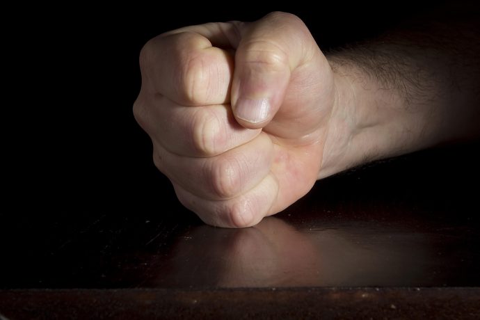 Eine Nahaufnahme einer geballten menschlichen Faust, die auf einer dunklen Holzoberfläche ruht, wobei der Unterarm vor einem schwarzen Hintergrund teilweise sichtbar ist - ein Symbol für Stärke und Entschlossenheit, das an Solanas unerschütterliche Entschlossenheit erinnert.