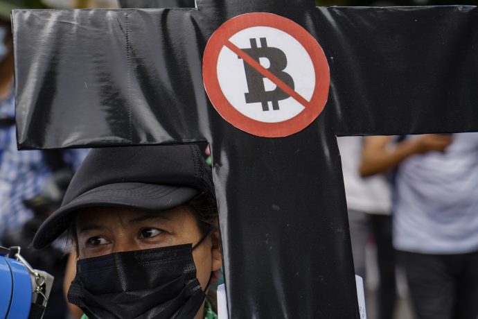 Eine Person mit schwarzer Maske hält ein schwarzes Kreuz mit einem rot durchgestrichenen Bitcoin-Symbol während einer NGO-Protestaktion, um auf die Bedenken über Bitcoin-Reserven und deren Vorteile für die Bevölkerung hinzuweisen.