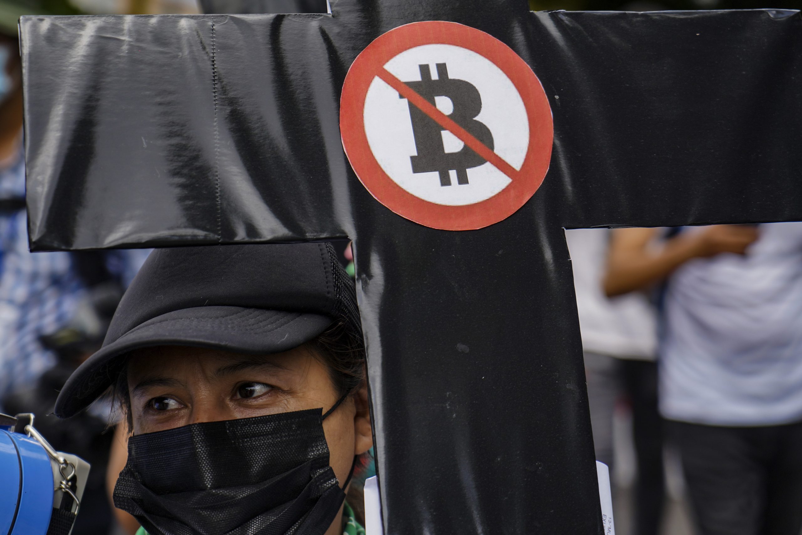 Eine Person mit schwarzer Maske hält ein schwarzes Kreuz mit einem rot durchgestrichenen Bitcoin-Symbol während einer NGO-Protestaktion, um auf die Bedenken über Bitcoin-Reserven und deren Vorteile für die Bevölkerung hinzuweisen.