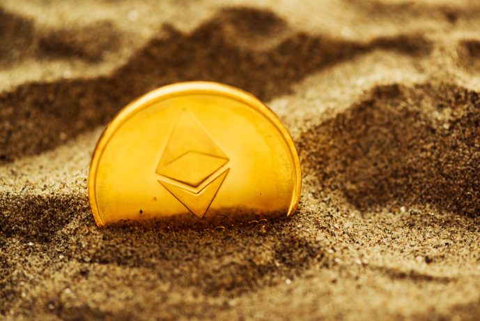 Eine goldene Ethereum-Münze mit dem unverwechselbaren Logo ist teilweise im Sand vergraben.