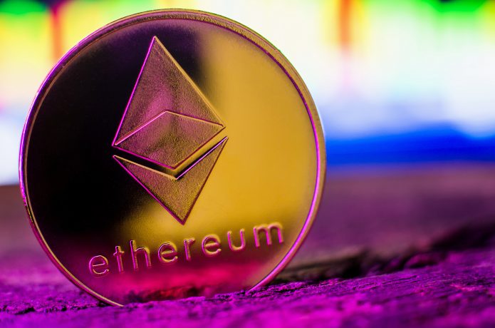 Eine goldfarbene physische Ethereum-Münze mit dem Ethereum-Logo und dem Wort "Ethereum" steht auf einer hölzernen Oberfläche mit einem unscharfen bunten Hintergrund.