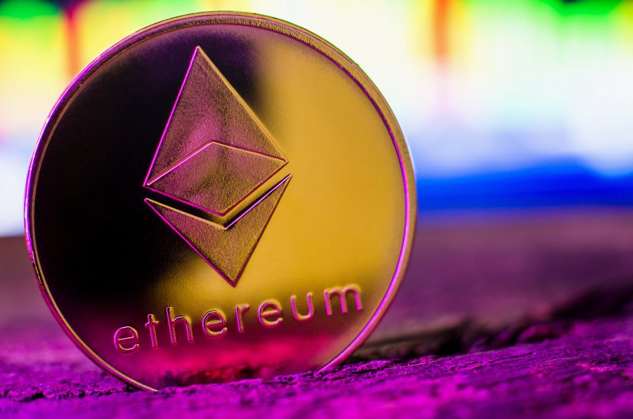 Eine goldfarbene physische Ethereum-Münze mit dem Ethereum-Logo und dem Wort "Ethereum" steht auf einer hölzernen Oberfläche mit einem unscharfen bunten Hintergrund.
