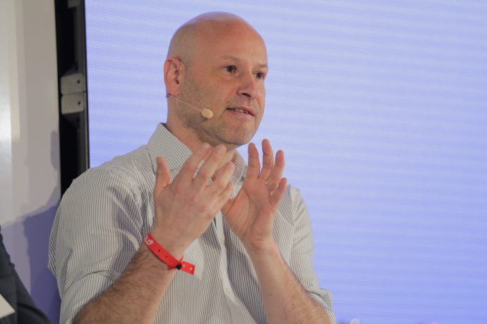 Ein Mann mit einem Mikrofon-Headset und einem roten Armband gestikuliert mit seinen Händen, während er vor einem hellen Hintergrund über Bitcoin spricht.