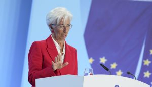 Eine Frau in einem roten Blazer spricht an einem Podium mit der Aufschrift EZB-Europäische Zentralbank, im Hintergrund ist eine EU-Flagge zu sehen.