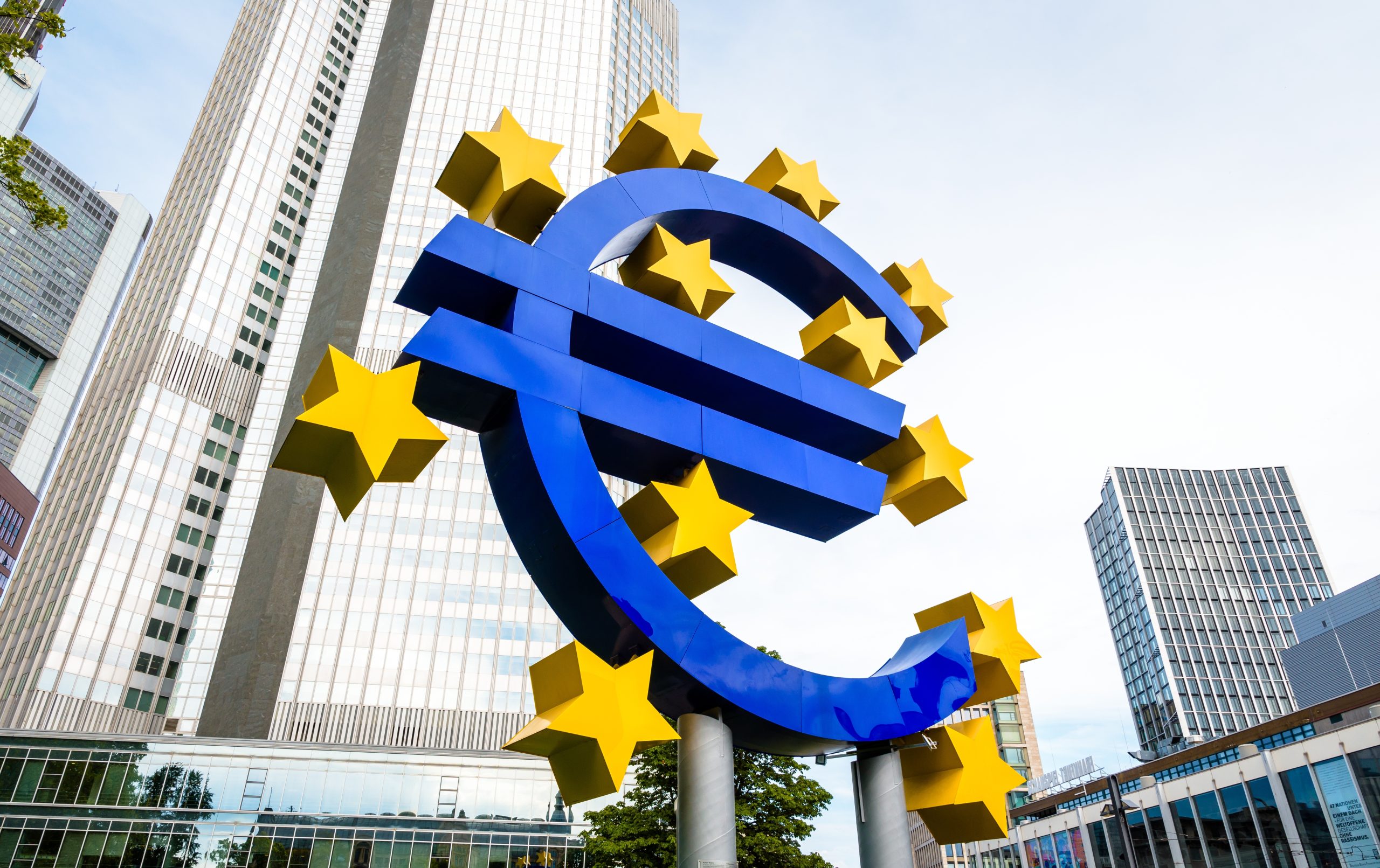 Eine große blaue Euro-Symbol-Skulptur mit gelben Sternen steht vor dem EZB-Hauptsitz inmitten moderner Bürogebäude in einem städtischen Gebiet.