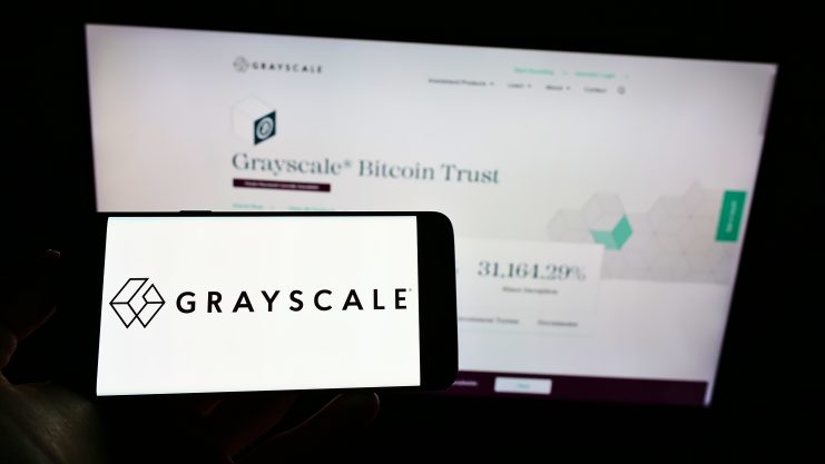 Ein Smartphone zeigt das Grayscale-Logo vor einem Computerbildschirm, auf dem die Grayscale Bitcoin Trust-Website zu sehen ist, was die Präsenz von Grayscale im digitalen Finanzwesen unterstreicht.