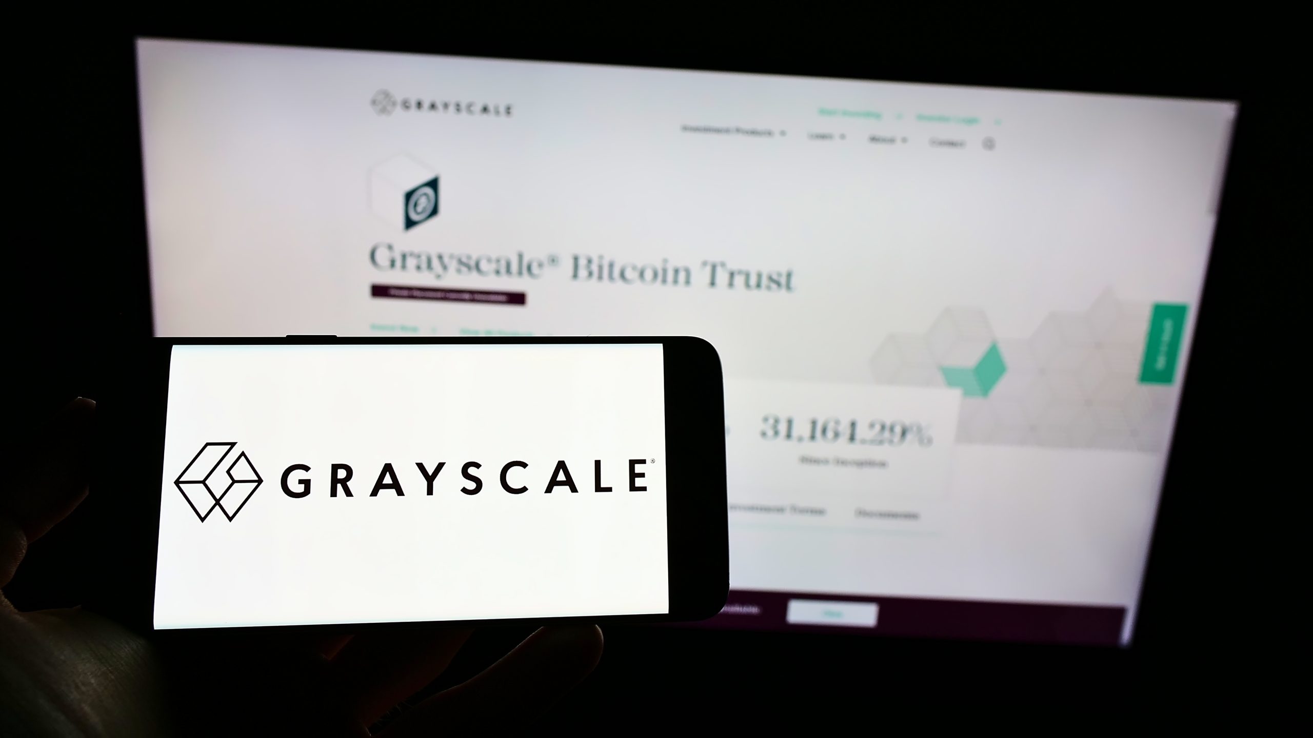 Ein Smartphone zeigt das Grayscale-Logo vor einem Computerbildschirm, auf dem die Grayscale Bitcoin Trust-Website zu sehen ist, was die Präsenz von Grayscale im digitalen Finanzwesen unterstreicht.