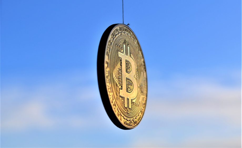 Ein goldfarbener Bitcoin-Token hängt an einer Schnur vor einem blauen Himmel mit ein paar Wolken und symbolisiert den Aufstieg des Bitcoin.