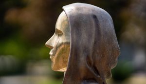 Seitliche Nahaufnahme einer Bronzestatue, die den Kopf und die Schultern einer Person mit Kapuze darstellt, inspiriert von Bitcoin, im Freien mit unscharfem Hintergrund.