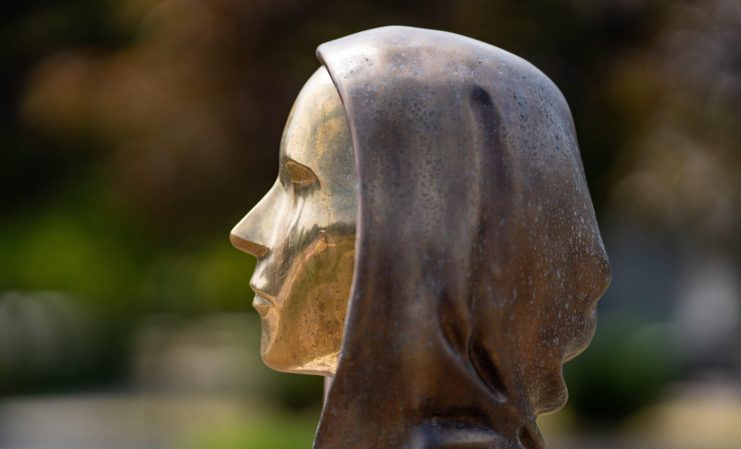 Seitliche Nahaufnahme einer Bronzestatue, die den Kopf und die Schultern einer Person mit Kapuze darstellt, inspiriert von Bitcoin, im Freien mit unscharfem Hintergrund.