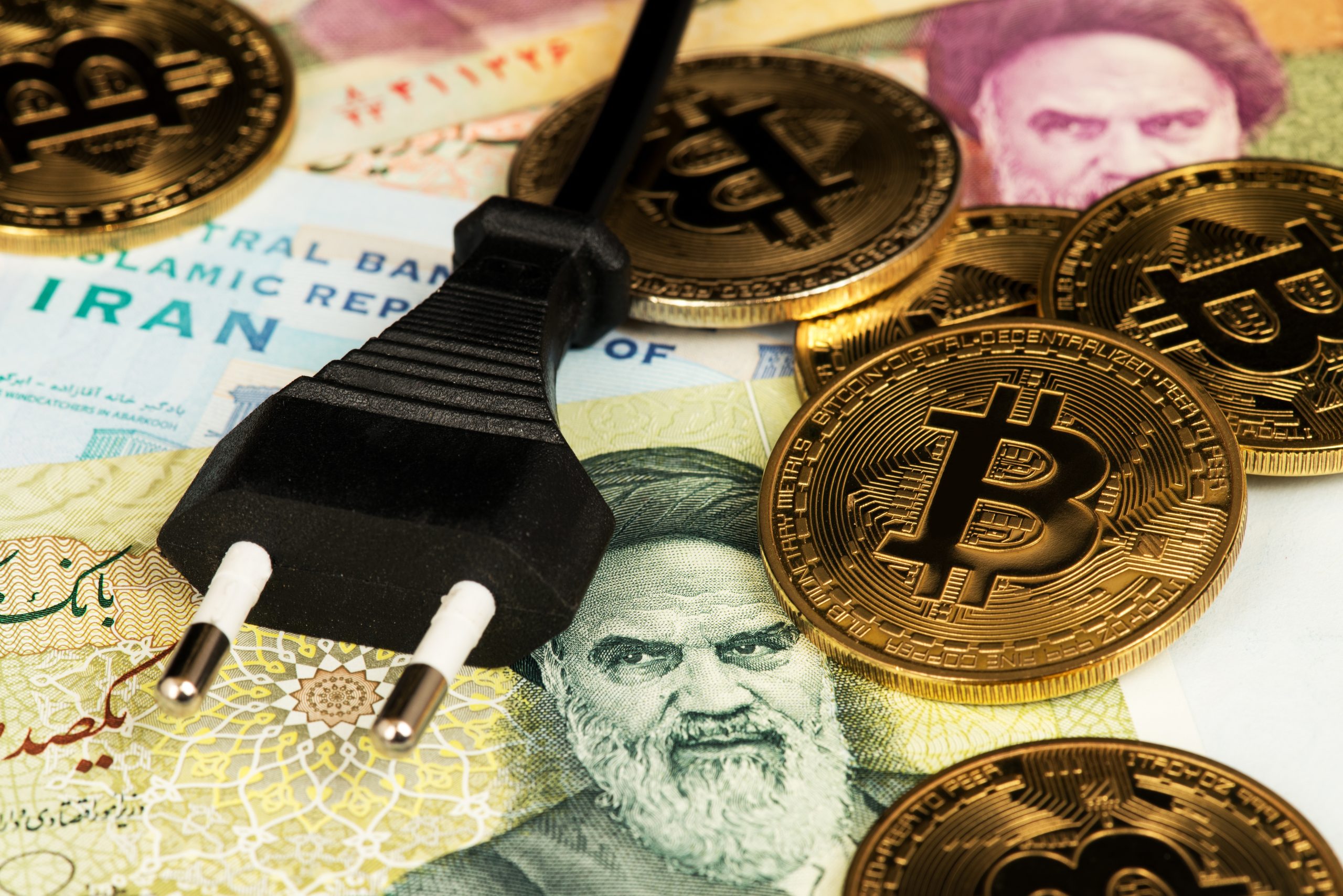 Ein schwarzer Stromstecker und mehrere goldene Bitcoin-Münzen liegen auf iranischen Banknoten mit Gesichtern, persischer Schrift und eindeutigen Symbolen des Iran.