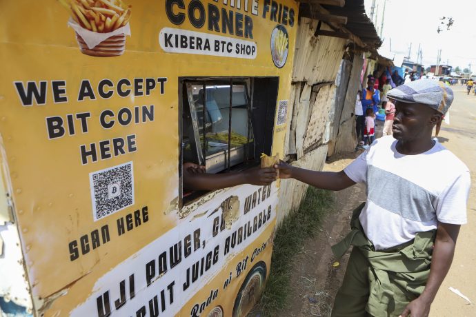Ein Mann zeigt den Daumen nach oben an einem Essensstand in Kibera, der stolz damit wirbt, dass er Bitcoin als Zahlungsmittel akzeptiert, wobei ein QR-Code und ein Bitcoin-Zeichen am Stand sichtbar sind.