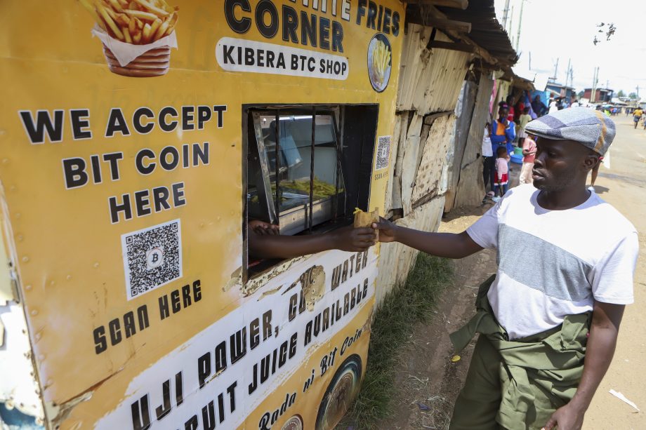 Ein Mann zeigt den Daumen nach oben an einem Essensstand in Kibera, der stolz damit wirbt, dass er Bitcoin als Zahlungsmittel akzeptiert, wobei ein QR-Code und ein Bitcoin-Zeichen am Stand sichtbar sind.