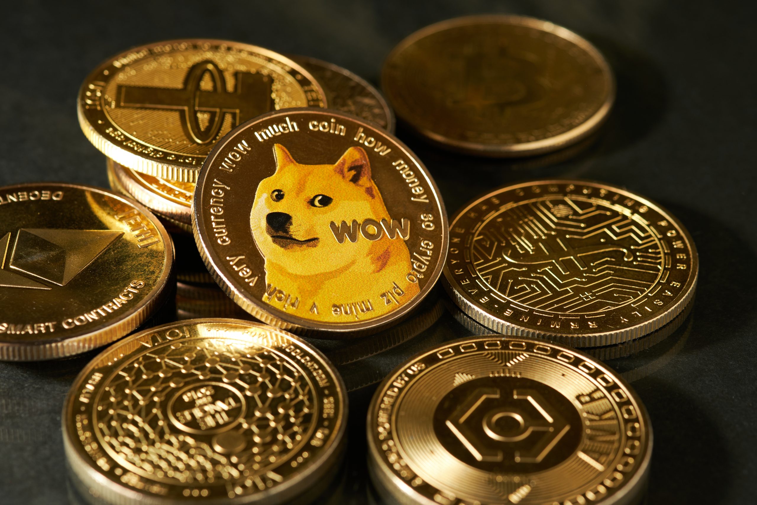 Mehrere physische Münzen, die verschiedene Kryptowährungen repräsentieren, sind auf einer Fläche verstreut, wobei in der Mitte eine Magnificent Seven-Auswahl von Token und ein Dogecoin mit dem Shiba Inu-Hund prominent dargestellt sind.