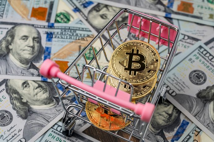 Ein kleiner Einkaufswagen mit glänzenden Bitcoin-Münzen steht auf einer Reihe von 100-Dollar-Scheinen.