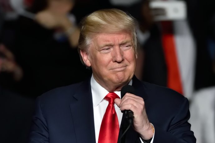 Ein Mann im Anzug und mit roter Krawatte hält ein Mikrofon in der Hand und steht vor einer verschwommenen Menge, die an Donald Trump erinnert, der zu seinen Anhängern spricht.