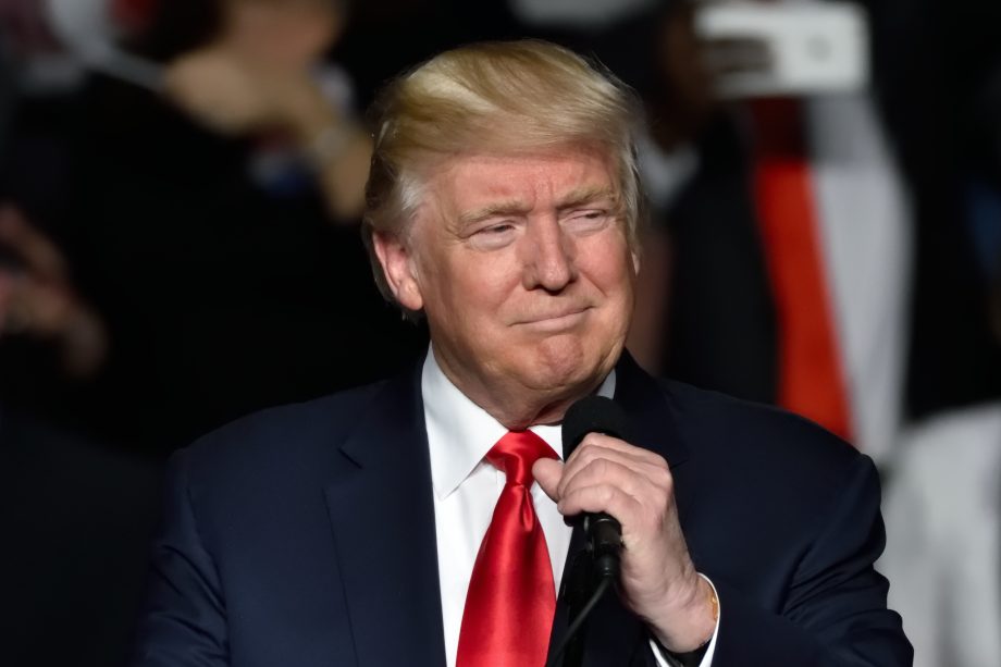 Ein Mann im Anzug und mit roter Krawatte hält ein Mikrofon in der Hand und steht vor einer verschwommenen Menge, die an Donald Trump erinnert, der zu seinen Anhängern spricht.