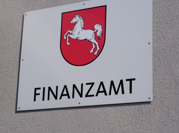 Ein Schild an der Wand zeigt ein rotes Schild mit einem weißen Pferd und dem Wort Finanzamt darunter, was auf die Rolle des Amtes bei der Überwachung der Krypto-Steuern hinweist.