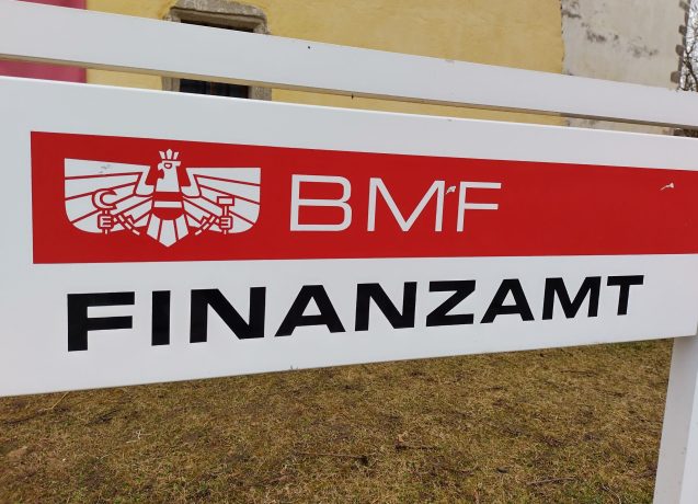 Ein Schild mit dem Logo des österreichischen Bundesministeriums für Finanzen (BMF) und dem Wort Finanzamt steht im Freien auf einer Wiese in der Nähe eines gelben Gebäudes und deutet auf die Aufsicht über Finanzangelegenheiten wie Steuern und die Regulierung von Stablecoins hin.
