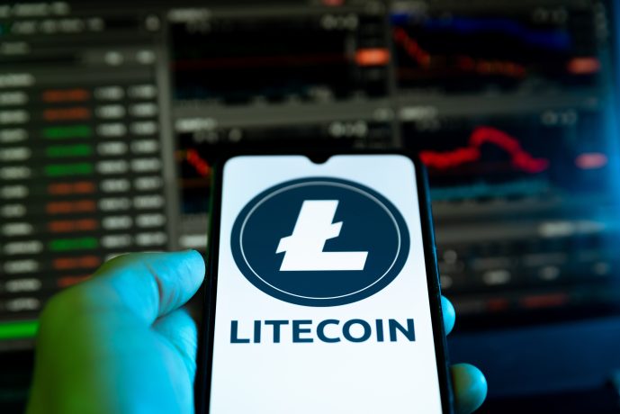 Eine Hand hält ein Smartphone, auf dem das Litecoin-Logo und der Text zu sehen sind, während auf den Bildschirmen im Hintergrund Kryptowährungs-Handelscharts und ein Krypto-Marktupdate zu sehen sind.