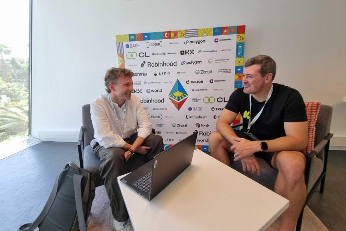 Zwei Männer sitzen sich auf Stühlen gegenüber und reden lächelnd über Ethereum. Zwischen ihnen steht ein Laptop auf einem Tisch und hinter ihnen ein Sponsor im Hintergrund.