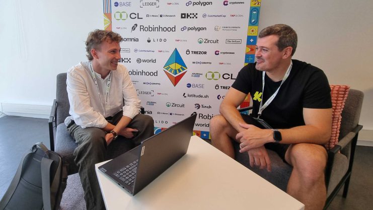 Zwei Männer sitzen sich auf Stühlen gegenüber und unterhalten sich mit einem Laptop auf dem Tisch zwischen ihnen; im Hintergrund befindet sich eine Sponsorentafel mit Logos von Technologieunternehmen, darunter Ethereum.
