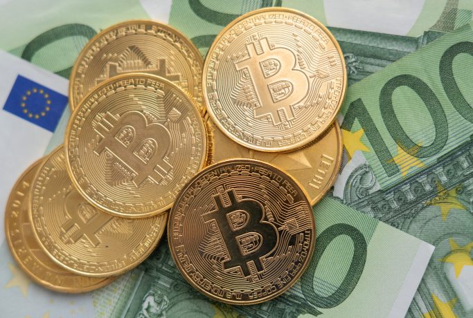 Mehrere goldfarbene Bitcoin-Token sind auf mehreren 100-Euro-Banknoten platziert, um die Überschneidung von traditioneller Währung und der digitalen Welt der Kryptowährung hervorzuheben.