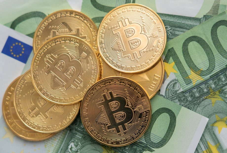 Mehrere goldfarbene Bitcoin-Token sind auf mehreren 100-Euro-Banknoten platziert, um die Überschneidung von traditioneller Währung und der digitalen Welt der Kryptowährung hervorzuheben.