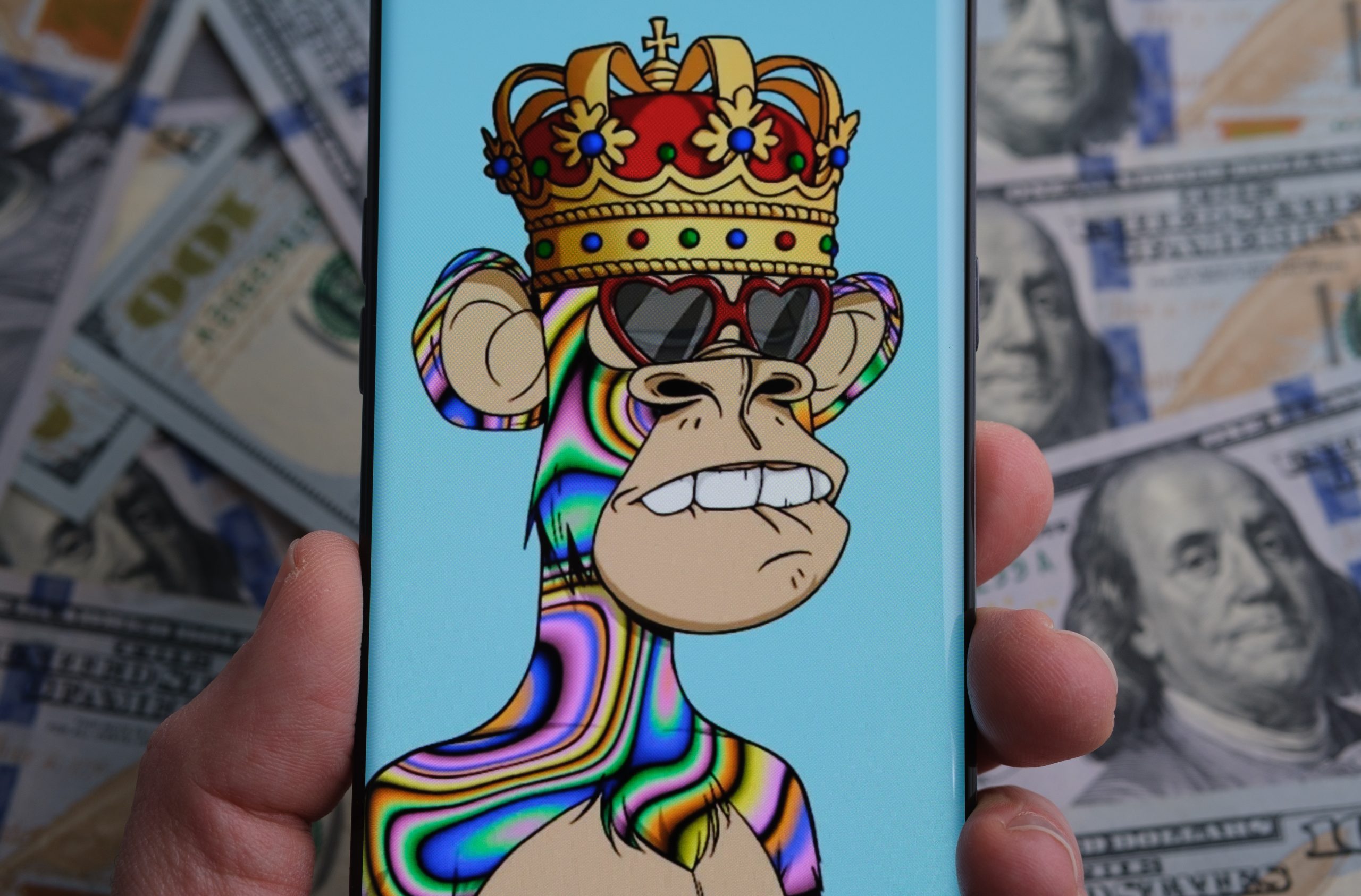 Eine Person hält ein Telefon in der Hand, auf dem ein Bored Ape-NFT mit Krone und herzförmiger Brille abgebildet ist. Im Hintergrund sind 100-US-Dollar-Scheine zu sehen, die den Rummel um NFTs und Memecoins verdeutlichen.