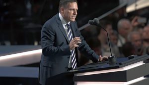 Peter Thiel, im Anzug, spricht auf einem Podium mit zwei Mikrofonen vor einem Publikum bei einer Indoor-Veranstaltung.