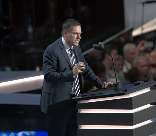 Peter Thiel, im Anzug, spricht auf einem Podium mit zwei Mikrofonen vor einem Publikum bei einer Indoor-Veranstaltung.