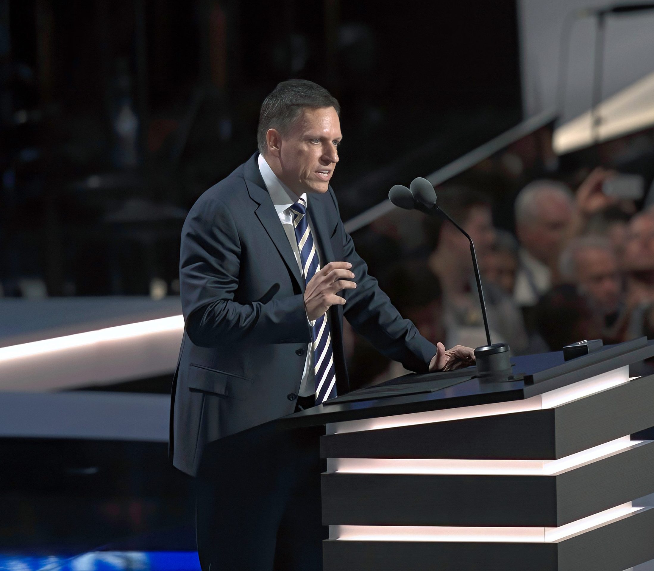 Peter Thiel, im Anzug, spricht auf einem Podium mit zwei Mikrofonen vor einem Publikum bei einer Indoor-Veranstaltung.