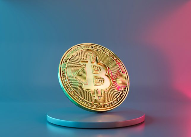 Eine goldene Bitcoin-Münze steht aufrecht auf einem runden Podest, ihre glänzende Oberfläche fängt das Licht vor einem Hintergrund mit blauem und rosa Farbverlauf ein.