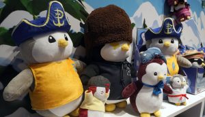 Eine Gruppe von Pudgy Penguins Plüschtieren in verschiedenen Kostümen, wie z. B. einem Piratenkostüm, einer Fliegerbrille und einem großen braunen Hut, steht in einem Regal vor einer verschneiten Bergkulisse.