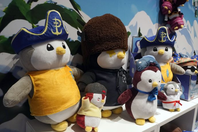 Eine Gruppe von Pudgy Penguins Plüschtieren in verschiedenen Kostümen, wie z. B. einem Piratenkostüm, einer Fliegerbrille und einem großen braunen Hut, steht in einem Regal vor einer verschneiten Bergkulisse.