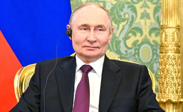 Ein Mann im Anzug sitzt auf einem goldenen Stuhl und trägt einen Ohrhörer. Hinter ihm ist ein gemusterter grüner Hintergrund und ein Teil einer blau-roten russischen Flagge zu sehen.