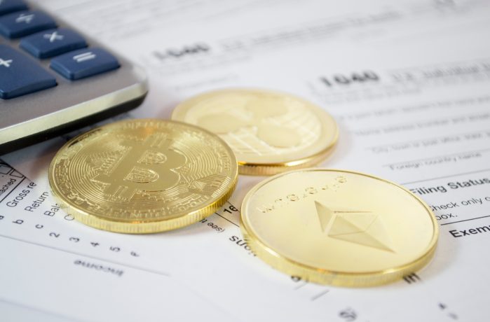 Drei goldfarbene Kryptowährungsmünzen (Bitcoin, Ethereum, Ripple) ruhen auf IRS-Steuerformularen, die auf die steuerlichen Auswirkungen des Einsatzes hinweisen, wobei in der Ecke ein Teil eines Taschenrechners zu sehen ist.