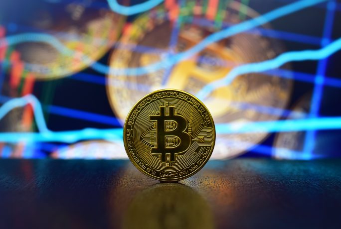 Eine goldfarbene Bitcoin-Münze steht aufrecht auf einer Fläche, vor einem verschwommenen Hintergrund mit Diagrammen und Linien, die Finanzmarktdaten anzeigen und auf die neuesten Bitcoin-Prognosetrends hinweisen.