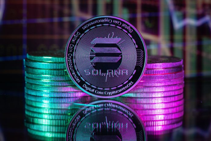 Eine Solana-Kryptowährungsmünze steht aufrecht vor gestapelten Münzen, die von grünen und rosafarbenen Lichteffekten mit einem digitalen Diagramm im Hintergrund beleuchtet werden und die lebendige Präsenz von Solana auf dem Kryptomarkt hervorheben.