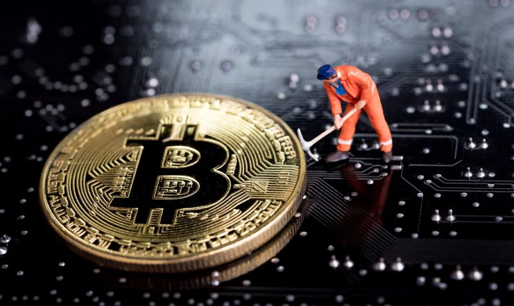 Eine Miniatur-Solo-Miner-Figur mit einer Spitzhacke steht neben einer großen Bitcoin-Goldmünze auf einem Computer-Motherboard.