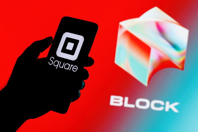 Eine Hand hält ein Smartphone, auf dem das Square-Logo abgebildet ist, während in der Ferne das Block-Logo und das Wort BLOCK auf rotem Hintergrund zu sehen sind, um die Präsenz von BLOCK unter den SP500-Unternehmen zu unterstreichen.