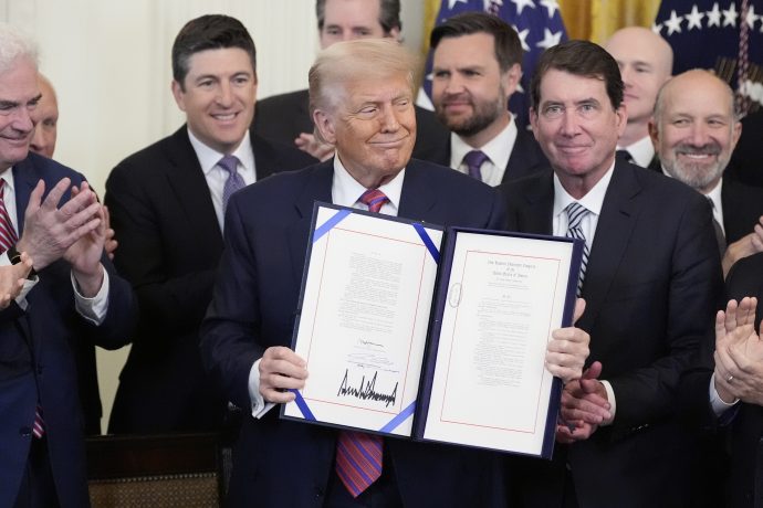 Donald Trump hält den unterzeichneten GENIUS-Act in die Höhe, während die Menschen um ihn herum in einem formellen Innenraum applaudieren.