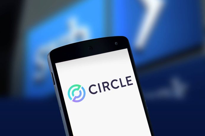 Ein Smartphone zeigt das Circle-Logo und den Namen auf dem Bildschirm an. Der blaue Hintergrund ist verschwommen und zeigt undeutliche Formen und Symbole, die die digitale Welt der Stablecoin-Lösungen widerspiegeln.